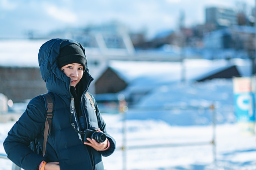 Winter snow travel concept, Happy traveler asian woman or photographer with backpack and camera relax in red brick warehouse at Hakodate city, Hokkaido, Japan