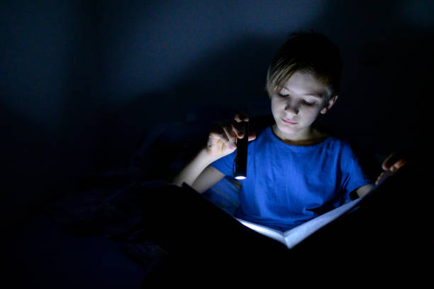 istock Teenage boy reading a book with flashlight in the bed 1199018083