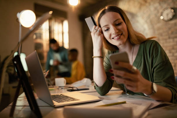 istock Happy freelancer holding credit card while using smart phone late in the office. 1199025956