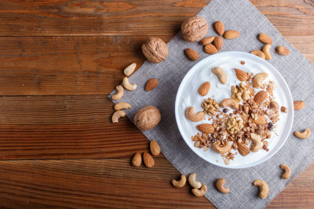 assiette blanche avec le yaourt grec, le granola, l'amande, la noix de cajou, les noix sur le fond en bois brun. vue du haut. copier l'espace. - yogurt greek culture milk healthy eating photos et images de collection