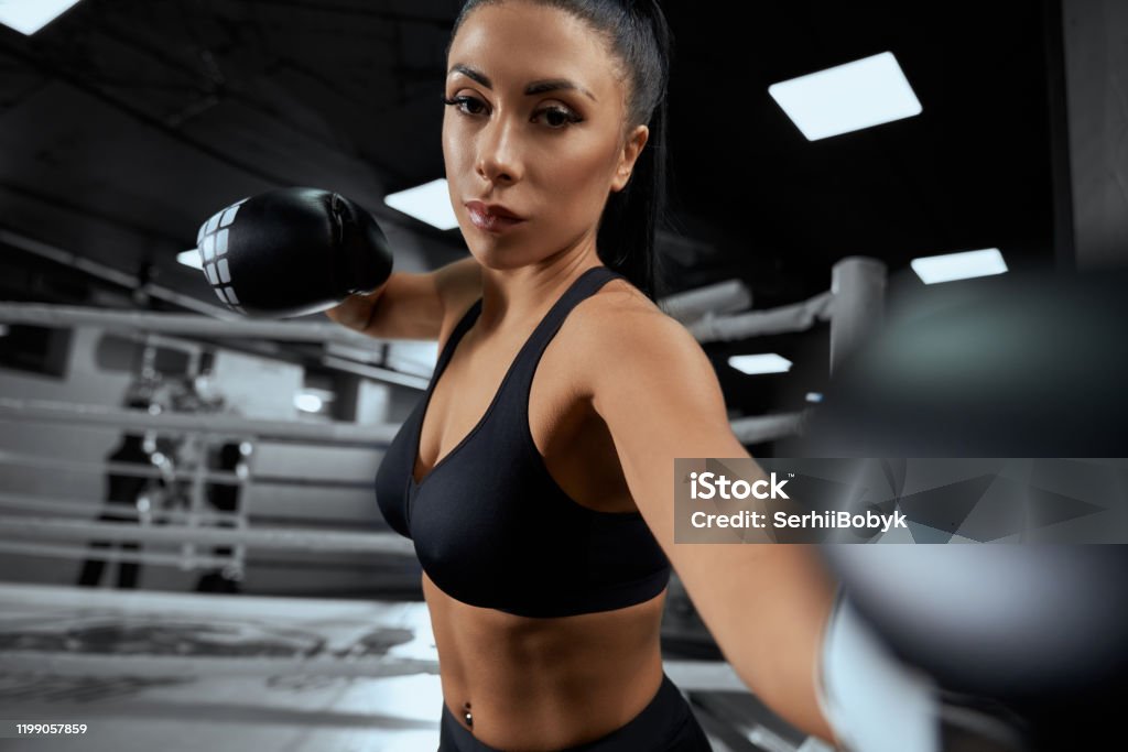 Woman fighting in boxing gloves. Front view of muscular woman with strong face wearing boxing gloves. Young attractive fighter with perfect make up looking at camera, posing and showing hit on boxing ring. Concept of sport, boxing. Boxing Ring Stock Photo