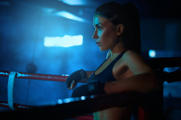 Side view of serious young professional female kickboxer wearing trendy sports outfit and bandages on hands. Fighter having rest after training, leaning on corner of ring. Concept of sport, boxing.