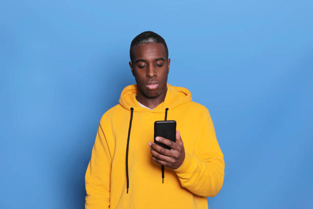 istock Cheerful African American man in yellow sweater using mobile phone application, happy hipster guy holding mobile phone on blue background 1199061843