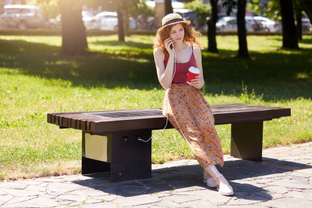 femme chargeant son téléphone sur le chargeur de panneau solaire incorporé dedans au banc de siège, buvant le café à emporter tout en parlant à quelqu'un par l'intermédiaire de son téléphone intelligent. technologie moderne, coconcept d'énergie  - cargill, incorporated photos et images de collection