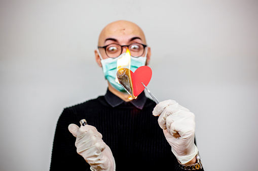 Bald guy with beard and eyeglasses dressed as doctor burning heart shaped paper with cigarette lighter