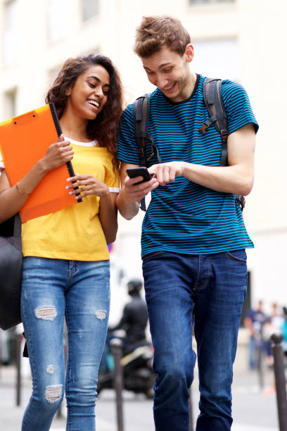smiling students looking at mobile phone while walking in city