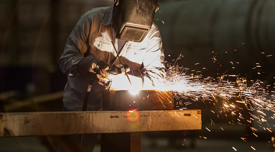 A mature man in his 50s working in a metal fabrication factory as a welder. He is covered in protective workwear, welding mask and gloves. Sparks are flying as he uses the welding torch.