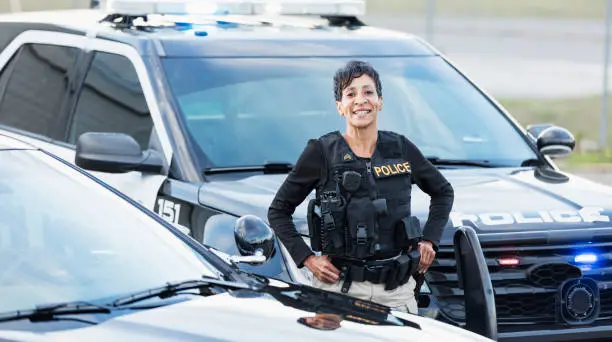 An African-American policewoman standing next to her patrol car, smiling confidently at the camera. She is a mature woman in her 40s, wearing a bulletproof vest and duty belt.