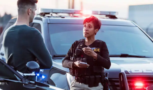 A policewoman taking a statement from a civilian outside her patrol car. The officer is a mature African-American woman in her 40s. She is talking with a young man in his 20s.