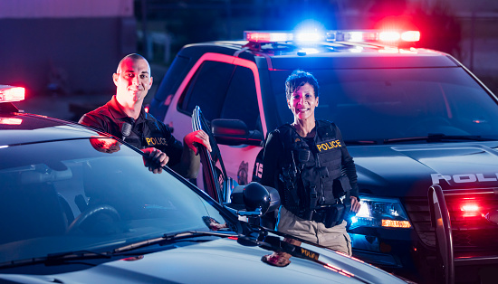 Two multi-ethnic police officers standing by the patrol cars, wearing bulletproof vests and duty belts. The policewoman is a mature African-American woman in her 40s. Her partner is a mid adult man in his 30s.
