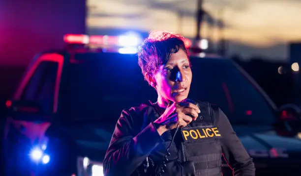 An African-American policewoman standing by her patrol car, talking on her radio with a serious expression on her face. She is a mature woman in her 40s, wearing a bulletproof vest and duty belt. It is nighttime and the emergency lights on her patrol car are flashing.
