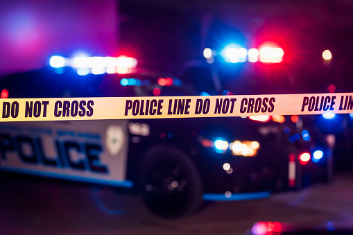 A crime scene at night. Police cars with emergency lights flashing behind yellow cordon tape declaring - police line, do not cross.