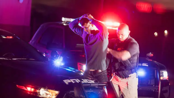 A police officer making an arrest, standing outside his patrol car at night, emergency lights flashing. The criminal is a young man in his 20s, standing his his hands on his head as the officer searches him.