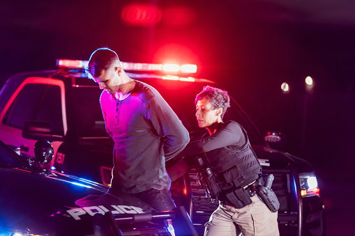 A policewoman putting handcuffs on a suspect. They are standing outside by a patrol car at night, emergency lights flashing behind them. The officer is a mature African-American woman in her 40s. The suspected criminal she is arresting is a young man in his 20s.