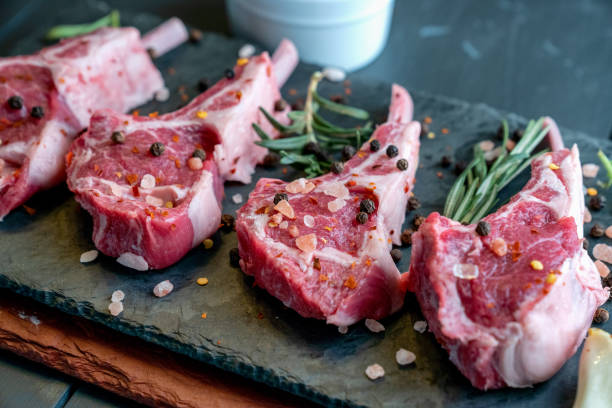 rohe lammkoteletts auf einem dunklen schiefer schneidebrett mit einem ramekin oder pesto an der seite mit rosmarin und anderen kräutern - lamm stock-fotos und bilder