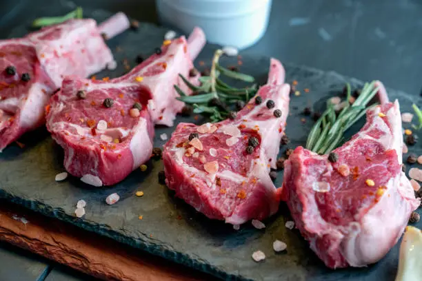 Photo of Raw Lamb Chops Prepared on a Dark Slate Cutting Board with a Ramekin or Pesto on the side with Rosemary and other herbs