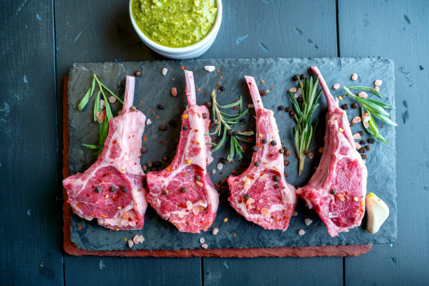 Raw Lamb Chops Prepared on a Dark Slate Cutting Board with a Ramekin or Pesto on the side with Rosemary and other herbs