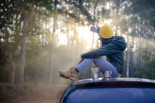 Woman traveller enjoy coffee time with taking shot selfie on her owns roof of the car with scenery view of the forest and light of mist morning in background