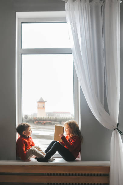 家の窓の上に座っている本を読んでいる子供たち。日の出の光で読む少年少女。 - window light window sill home interior ストックフォトと画像
