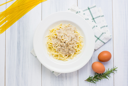 Delicious spaghetti carbonara with white sauce with tablecloth on white wood plank table top with raw food material at background.