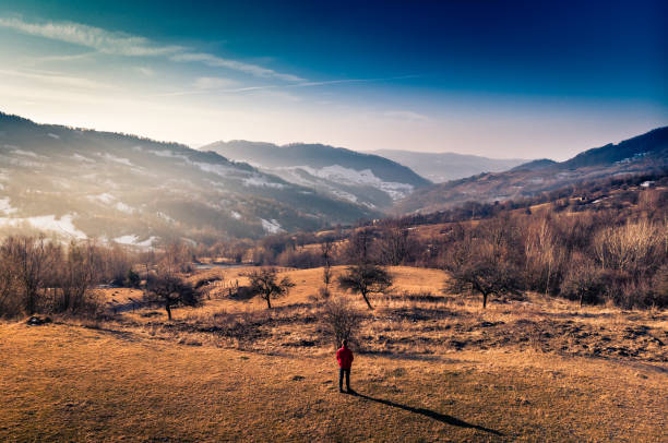 Aerial color image taken by drone, depicting a man wearing a red jacket standing alone in a barren winter landscape in the heart of the Carpathian mountains in Transylvania, Romania. The man is presented in rear view and is unrecognisable. In the distance are the rolling foothills of the mountains with patches of snow, and a dazzling blue sky. Room for copy space.