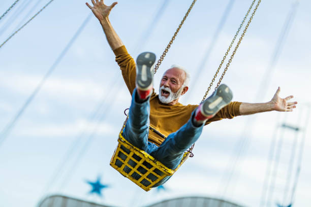 Carefree mature man having fun on chain swing ride in amusement park. Happy senior man having fun while riding on chain swing at amusement park and looking at camera. young at heart stock pictures, royalty-free photos & images