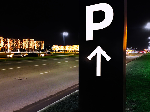 Tall black and white PARKING sign with an up arrow on the side of road at night. Winter trees decorated with light on the background