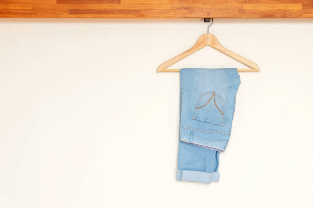 Light blue denim pants hanging on wood hanger on minimal open closet storage with copy space on wall background. stock photo