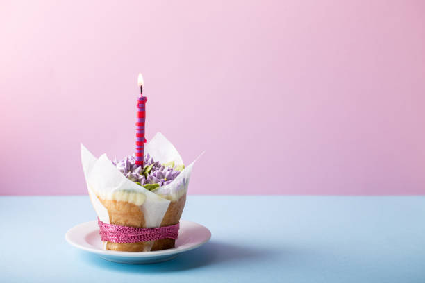 istock Flower cupcake  with single candle on blue  table on pink background. Beautiful sponge cup cakes decorated with buttercream lilac flower. Copy space. 1199166549