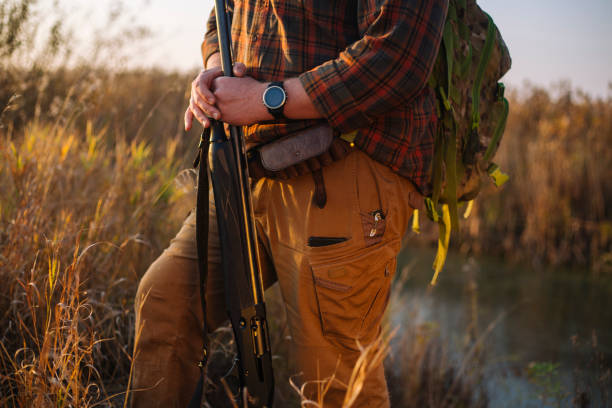 las manos de hunter sosteniendo un arma. hombre de pie al aire libre cerca del río y usando camisa a cuadros, pantalones de camello, cinturón de munición de cuero y reloj inteligente. foto con enfoque selectivo - bullet belt ammunition cartridge fotografías e imágenes de stock