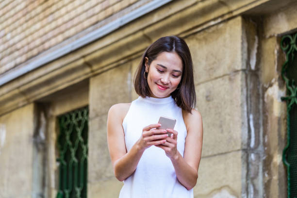 Portrait of a young, attractive and beautiful Asian woman smiling as she texting on her smartphone int he city. She is looking away as she talks animatedly and happily on her mobile device. retrato de una joven, atractiva y hermosa mujer asiática sonriendo mientras le envía mensajes de texto en su teléfono inteligente en la ciudad. ella está mirando hacia otro lado mientras habla animada y felizmente en su dispositivo móvil.