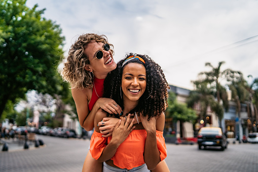 Two young beautiful smiling Argentinian women having fun at city street, having a piggyback ride. Shooting Buenos Aires, Argentina.
