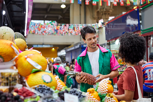 Confident male vendor selling pineapple to young woman. Smiling man is assisting female customer in choosing fruits. They are standing at market stall.