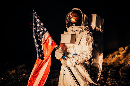 One man, astronaut exploring the land on the other planet alone at night, holding a USA national flag.