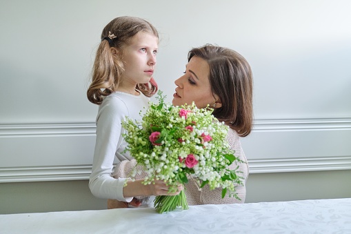 Talking mother and little daughter, unhappy child with tearful eyes looking out the window, woman hugged girl and reassures. In hands bouquet of flowers presented on Mothers Day