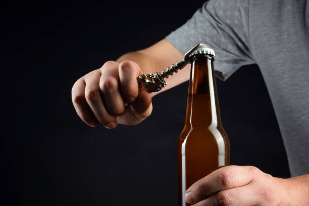 istock Men opening cold bottle of beer with cap on black background. Hands cracking refrigerated wheat or lager beer with an opener on dark background 1199195211