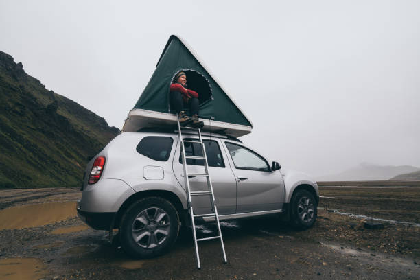 Young woman sitting on a ladder next to offroad car with tent on the roof in Landmannalaugar, Iceland stock photo