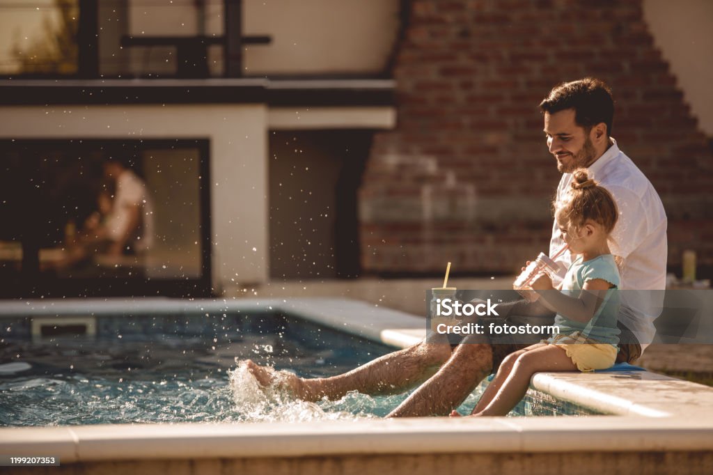 Father and daughter funny time by the pool Side view of lovely scene where daughter and dad plunge their feet in a pool while drinking fresh juices. Family Stock Photo