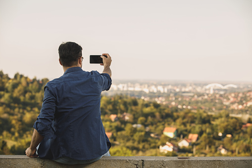 Rear view of man in casual clothes taking pictures with his phone in a lookout location with beautiful view,