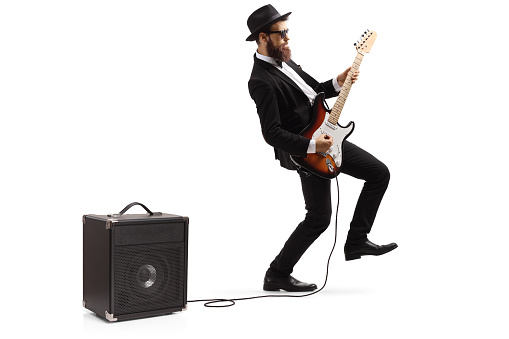 Full length shot of a bearded male artist in a suit playing a plugged electric guitar isolated on white background