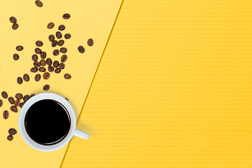 A cup of black coffee and scatted coffee beans on yellow background, top view. Monday coffee