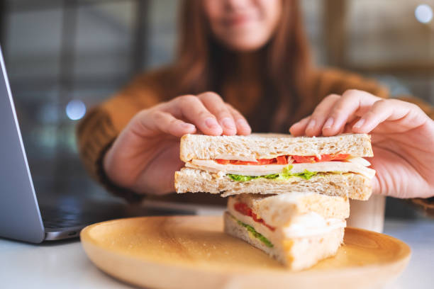 eine frau hält und isst vollkorn-sandwich während der arbeit am laptop-computer - sandwich healthy eating bread snack stock-fotos und bilder