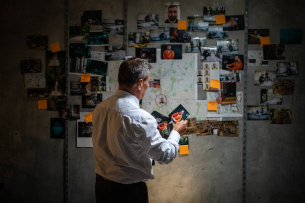 Mature police detective looking at pictures while standing in front of the wall Rear view of gray-haired police detective looking pictures in front of the wall with map, pictures and adhesive notes on it, and searching for a lead on his case wall of monitors stock pictures, royalty-free photos & images