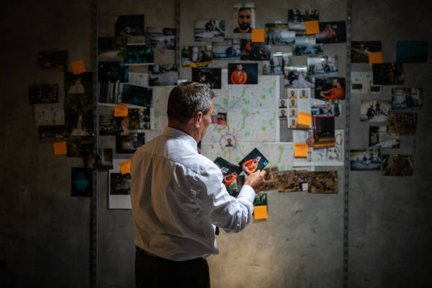 Mature police detective looking at pictures while standing in front of the wall Rear view of gray-haired police detective looking pictures in front of the wall with map, pictures and adhesive notes on it, and searching for a lead on his case criminal investigation photos stock pictures, royalty-free photos & images