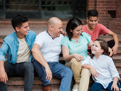 A happy latin family with three children sitting on the steps outside and smiling at the little girl.