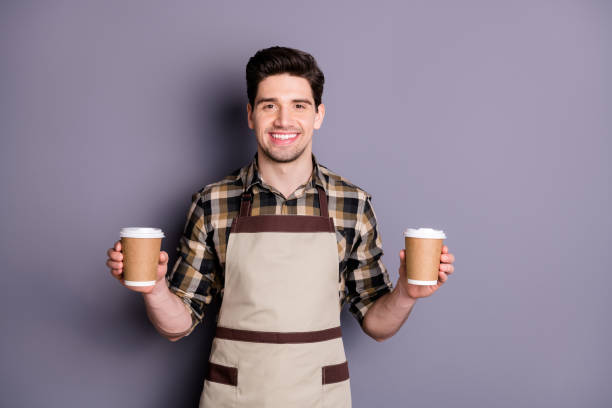 istock Photo of attractive worker guy holding hands two paper coffee cups inviting visitors try aroma coffee professional barista wear apron plaid shirt isolated grey color background 1199248421