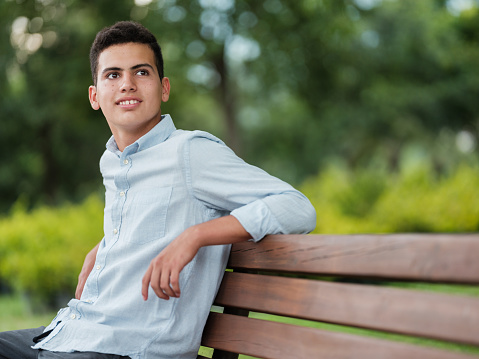 A handsome latin teenage boy sitting on a bench at the park and looking away.