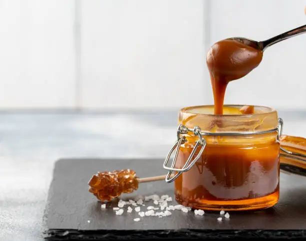 Spoon with tasty caramel sauce over jar on table.