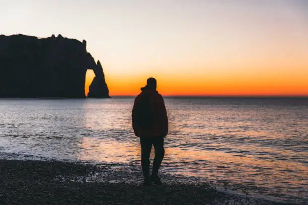 Young man walking at the beautiful northern beach with waves and enjoying the view of big natural arch and cliffs in Etretat, Normandy, Northern France