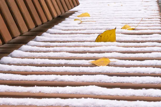 Wooden bench seat covered with white snow and autumn leaves. Selective focus.